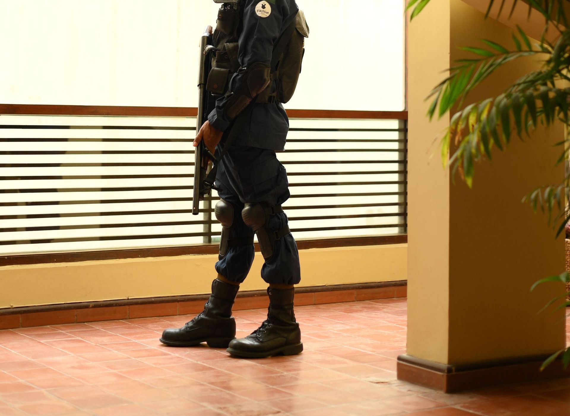 A security guard in uniform stands indoors, observing through a window by a staircase.