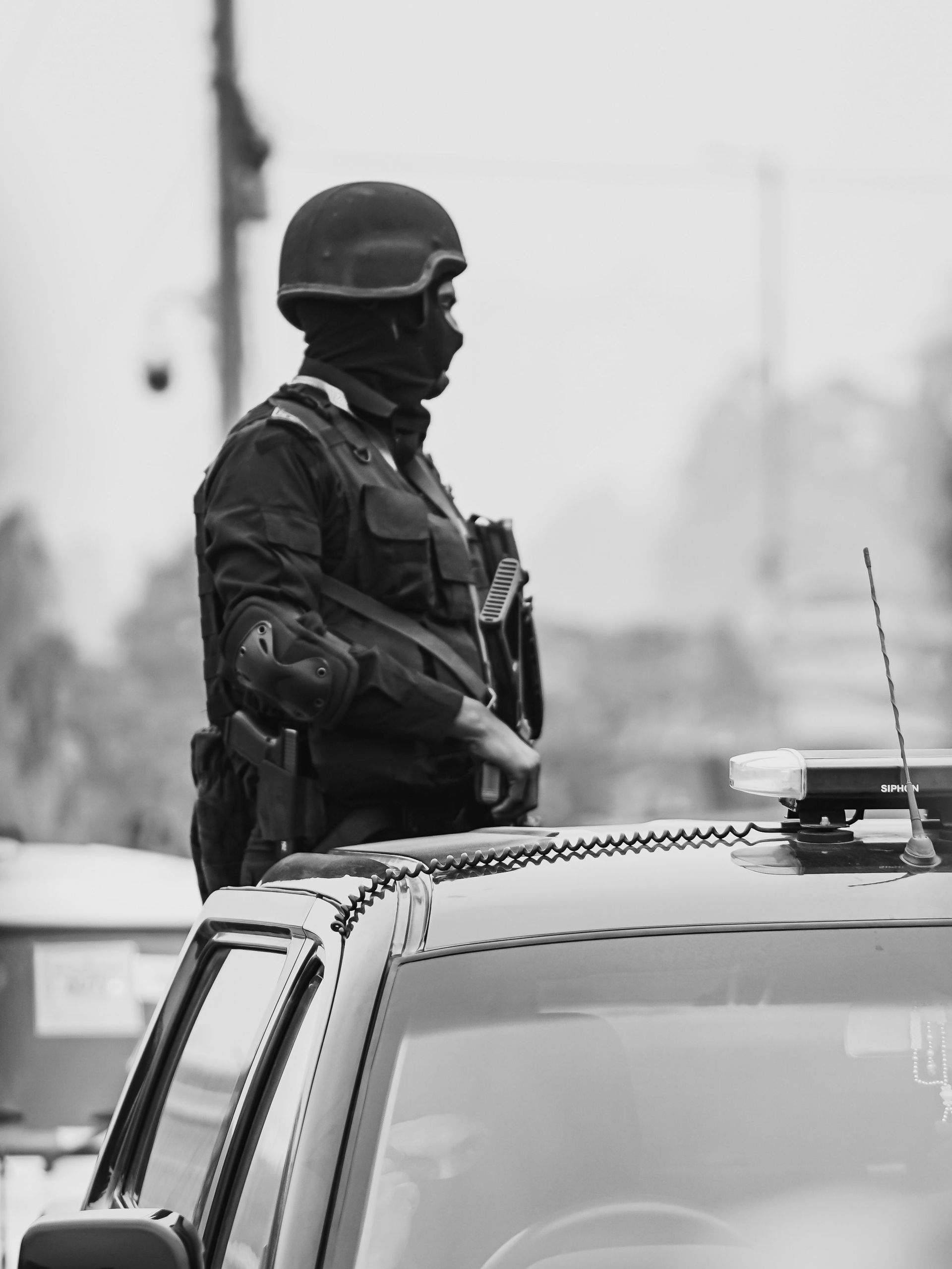 Black and white photo of armed soldier on duty in New Delhi street, standing on a vehicle.